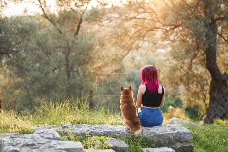 A woman with pink hair enjoys a serene moment in nature, sharing a gentle touch with a Shiba Inu dog on a stone in a lush grove