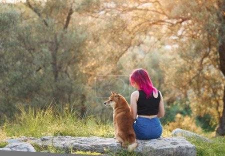 Una mujer con el pelo rosa disfruta de un momento sereno en la naturaleza, compartiendo un toque suave con un perro Shiba Inu en una piedra en un exuberante bosque