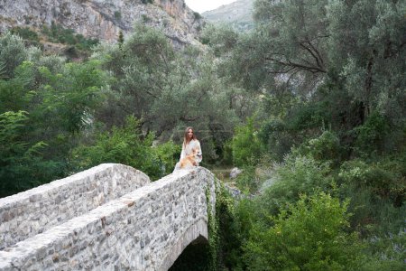 A woman in a flowing dress enjoys a serene moment with her dog on an ancient stone bridge in a lush valley. 