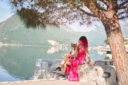 Una mujer con el pelo rosa se sienta al lado de un perro Shiba Inu, ambos mirando hacia un lago tranquilo. La escena captura un momento sereno de compañía en la naturaleza