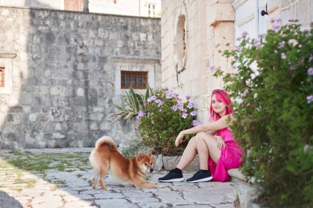 Photo for A woman with vibrant pink hair gently pets a Shiba Inu dog in a quaint stone-paved alley, a tender moment of connection. - Royalty Free Image
