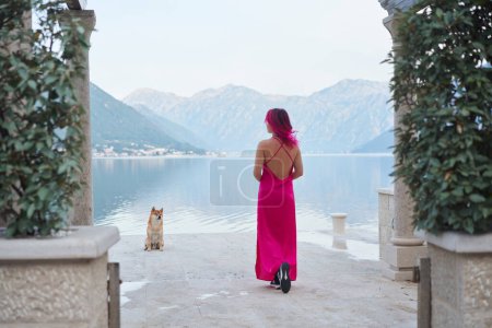 Una mujer con el pelo rosa en un vestido que fluye se levanta frente a una vista impresionante junto al lago, con un Shiba Inu mirando hacia ella