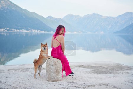 Eine Frau mit rosafarbenen Haaren teilt einen Moment am See mit einem Shiba Inu, dem Hund auf einem Stein, während beide über das Wasser schauen. Die bergige Landschaft spiegelt sich im stillen See