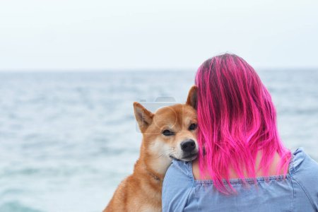 Ein Shiba-Inu-Hund schmiegt sich gemütlich an eine Frau mit auffallend rosafarbenem Haar und blickt auf das ruhige Meer. Ihre liebevolle Umarmung verkörpert eine besondere Mensch-Tier-Bindung
