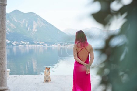 Une femme aux cheveux roses dans une robe fluide se tient face à une vue imprenable sur le lac, avec un Shiba Inu regardant vers elle