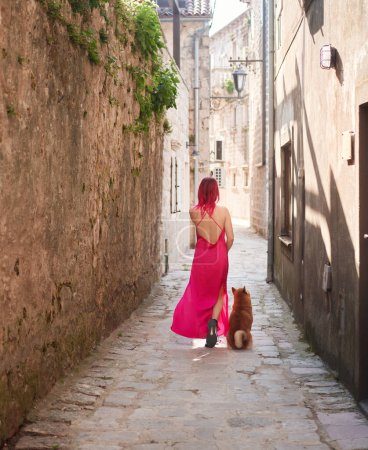 A woman in a striking pink dress strolls down an ancient cobblestone lane with her Shiba Inu dog, both moving away into the distance. 