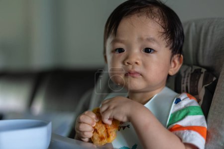 Téléchargez les photos : Adorable joyeux et heureux bébé chinois de 1ans ayant de la nourriture tout en s'asseyant sur une chaise bébé - en image libre de droit