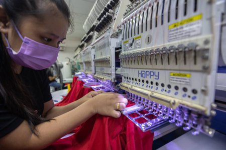 Foto de Kota Kinabalu, Sabah, Malasia 24 de agosto. 2021: Una trabajadora con mascarilla médica opera la máquina de bordado en la industria textil en la fabricación de prendas de vestir - Imagen libre de derechos