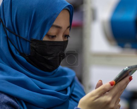 Photo for Asian muslim woman wearing medical face mask for prevent dust and infection virus, using smartphone during office hour - Royalty Free Image