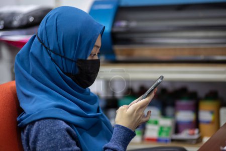 Téléchargez les photos : Femme musulmane asiatique portant un masque médical pour prévenir la poussière et le virus de l'infection, en utilisant un smartphone pendant l'heure de bureau - en image libre de droit