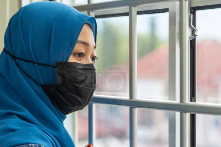 Téléchargez les photos : Une fille musulmane regardant par la fenêtre et portant un masque de protection et de récupération de la maladie à la maison. En quarantaine. Patient isolé pour prévenir l'infection. Une pandémie. Maison. - en image libre de droit