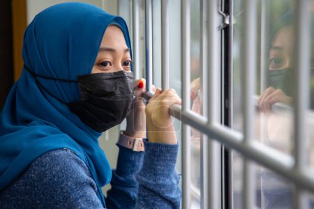 Foto de Una chica musulmana mirando a través de la ventana y usando máscara de protección y recuperación de la enfermedad en el hogar. Cuarentena. Paciente aislado para prevenir infección. Pandemia. Casa. - Imagen libre de derechos