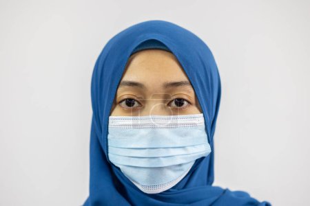 Téléchargez les photos : Gros plan portrait d'attrayant contenu sérieux muslimah portant bleu pastel hijab masque blanc isolé sur fond blanc - en image libre de droit