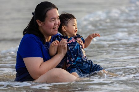 Asiática china madre con feliz 1 a 2 años de edad niño disfrutando jugando en la playa con salpicaduras de agua de mar caliente