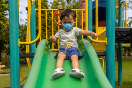 Téléchargez les photos : Asiatique enfant jouer au parc aire de jeux avec masque médical pendant la journée - en image libre de droit