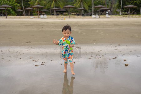 Foto de Feliz niño de 1-2 años disfrutando jugando en la playa con agua tibia salpicada - Imagen libre de derechos