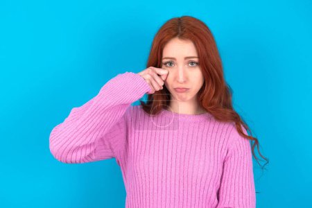 Photo for Unhappy red haired woman wearing pink sweater over blue background crying while posing at camera whipping tears with hand. - Royalty Free Image