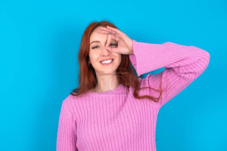 Foto de Mujer pelirroja vistiendo suéter rosa sobre fondo azul con la cara feliz sonriendo haciendo signo bien con la mano en el ojo mirando a través del dedo. - Imagen libre de derechos