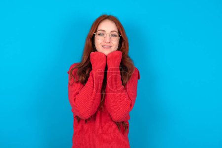 Foto de Mujer joven satisfecha vistiendo suéter rojo sobre fondo azul toca la barbilla con ambas manos, sonríe agradablemente, se regocija buen día con amante - Imagen libre de derechos