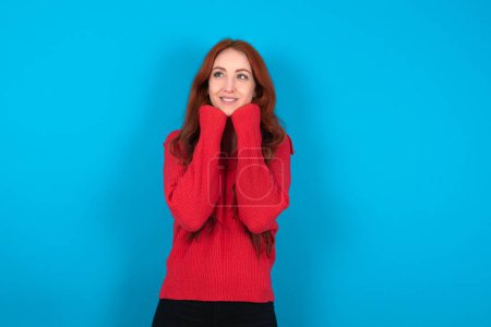 Foto de Joven mujer vistiendo suéter rojo sobre fondo azul sostiene las manos bajo la barbilla, contento de escuchar palabras conmovedoras de extraño - Imagen libre de derechos