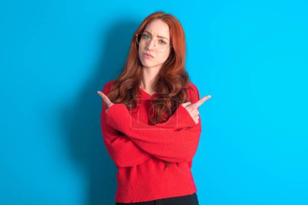 Foto de Mujer joven seria vistiendo suéter rojo sobre fondo azul cruza las manos y puntos en diferentes lados duda entre dos artículos. Concepto de decisión difícil - Imagen libre de derechos