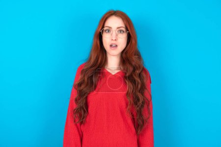 Foto de Mujer joven impactada con suéter rojo sobre fondo azul mira fijamente ojos con micrófonos mantiene la boca abierta ha sorprendido a la expresión. Concepto de Omg - Imagen libre de derechos