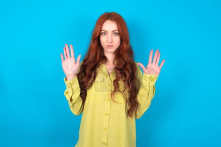 Photo for Serious beautiful red haired woman wearing green shirt over blue studio background pulls palms towards camera, makes stop gesture, asks to control your emotions and not be nervous - Royalty Free Image
