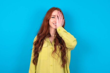 Téléchargez les photos : Jeune femme rousse caucasienne portant une chemise verte sur fond bleu couvrant un œil avec sa main, sourire confiant sur le visage et émotion surprise. - en image libre de droit