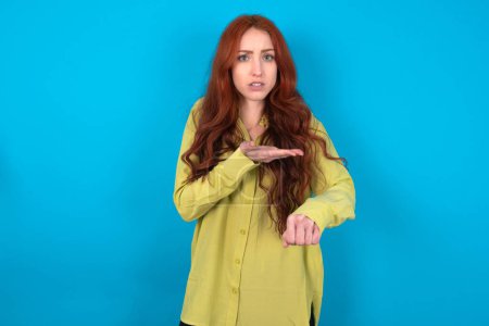 Téléchargez les photos : Jeune caucasienne rousse femme portant chemise verte sur fond bleu pressé pointant vers regarder le temps, impatience, bouleversé et en colère pour retard de délai. - en image libre de droit