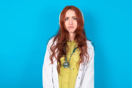 Photo for Serious displeased Young redhead caucasian doctor woman over blue background looks puzzled at camera being angry wears stereo headphones listens music while walking at street - Royalty Free Image