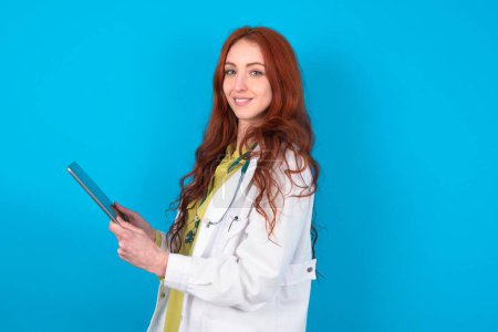 Téléchargez les photos : Photo optimiste Jeune rousse caucasienne médecin femme sur fond bleu tenir comprimé - en image libre de droit