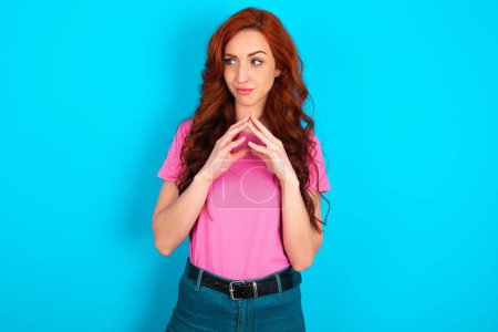 Photo for Young caucasian redhead woman wearing pink t-shirt over blue background steepled fingers and looks mysterious aside has great evil plan in mind - Royalty Free Image