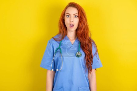 Schockierte junge rothaarige Ärztin mit gelbem Studiohintergrund starrt verdutzte Augen an und hält den Mund offen. Omg-Konzept