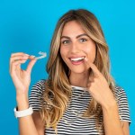 Young beautiful blonde woman wearing striped t-shirt over blue studio background holding an invisible aligner and pointing to her perfect straight teeth. Dental healthcare and confidence concept.