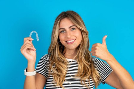 Young beautiful blonde woman wearing striped t-shirt over blue studio background holding an invisible braces aligner and rising thumb up, recommending this new treatment. Dental healthcare concept.