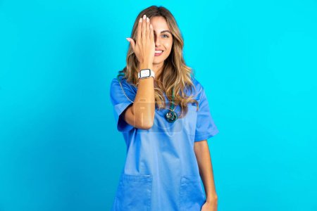 Téléchargez les photos : Jeune belle médecin femme debout sur fond bleu studio couvrant un oeil avec sa main, sourire confiant sur le visage et l'émotion surprise. - en image libre de droit