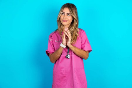 Photo for Young beautiful doctor woman standing over blue studio background steepled fingers and looks mysterious aside has great evil plan in mind - Royalty Free Image
