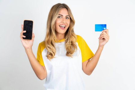 Photo for Young beautiful woman wearing football T-shirt over white background opened bank account, holding smartphone and credit card, smiling, recommend use online shopping application - Royalty Free Image