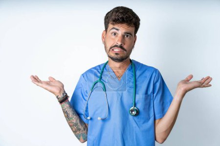Photo for Clueless handsome nurse man wearing surgeon uniform over white background shrugs shoulders with hesitation, faces doubtful situation, spreads palms, Hard decision - Royalty Free Image