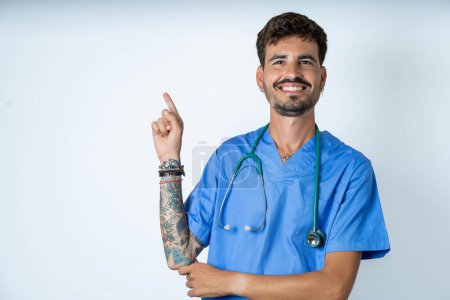 Téléchargez les photos : Belle infirmière souriante homme portant uniforme de chirurgien sur fond blanc indiquant doigt espace vide montrant les meilleurs prix bas - en image libre de droit