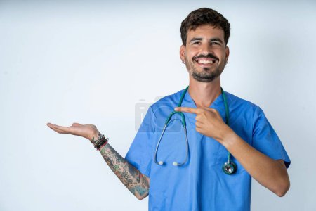 Foto de Divertido enfermero guapo vistiendo uniforme de cirujano sobre fondo blanco sosteniendo palma abierta nuevo producto. Quiero comprarlo.! - Imagen libre de derechos