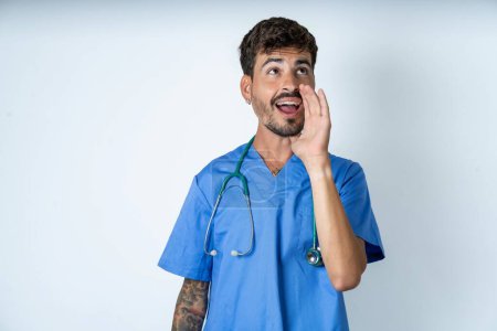 Foto de Guapo enfermera hombre vistiendo cirujano uniforme sobre blanco fondo escuchar increíble privado noticias impresionado grito compartir - Imagen libre de derechos