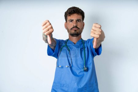 Photo for Handsome nurse man wearing surgeon uniform over white background being upset showing thumb down with two hands. Dislike concept. - Royalty Free Image