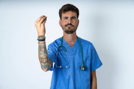 Foto de Guapo enfermero vistiendo uniforme de cirujano sobre fondo blanco gesto enojado típico italiano gesto con la mano, mirando a la cámara - Imagen libre de derechos