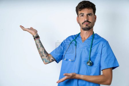 Téléchargez les photos : Belle infirmière homme portant uniforme de chirurgien sur fond blanc pointant de côté avec les deux mains montrant quelque chose d'étrange et disant : Je ne sais pas ce que c'est. Concept de publicité. - en image libre de droit