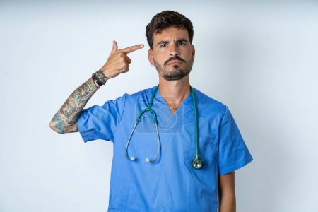 Photo for Unhappy handsome nurse man wearing surgeon uniform over white background makes suicide gesture and imitates gun with hand, curves lips keeps two fingers on temple, shoots, being tired of everything, - Royalty Free Image