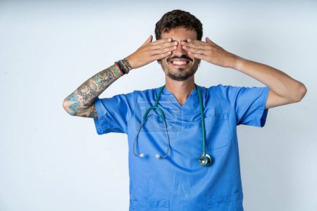 Photo for Handsome nurse man wearing surgeon uniform over white background covering eyes with hands smiling cheerful and funny. Blind concept. - Royalty Free Image