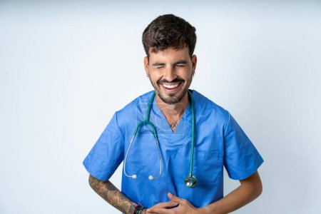 Foto de Guapo enfermero vistiendo uniforme de cirujano sobre fondo blanco sonriendo y riendo en voz alta porque divertido chiste loco con las manos en el cuerpo. - Imagen libre de derechos