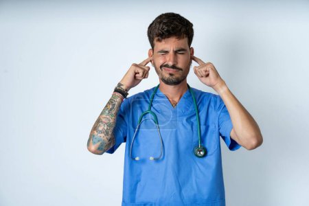Photo for Handsome nurse man wearing surgeon uniform over white background covering ears with fingers with annoyed expression for the noise of loud music. Deaf concept. - Royalty Free Image