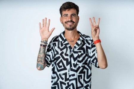 Foto de Hombre con tatuaje vistiendo camisa de verano de pie sobre fondo blanco aislado mostrando y apuntando hacia arriba con los dedos número ocho mientras sonríe confiado y feliz. - Imagen libre de derechos